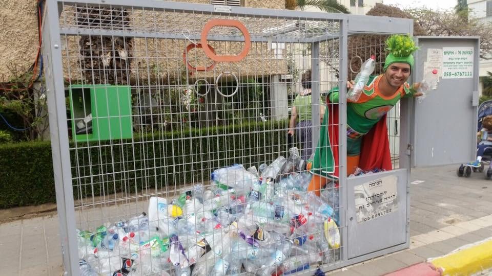 אביעד בראשי בדמות האיש הירוק, עומד בתוך כלוב מיחזור מלא בקבוקי פלסטיק